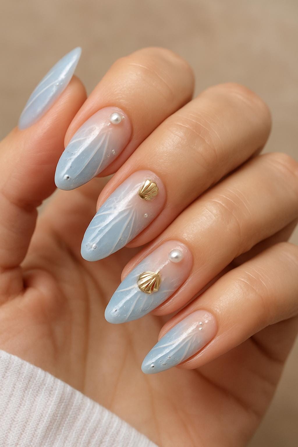 Almond nails with soft blue ombré, white feathered lines, pearls, and gold seashell accents